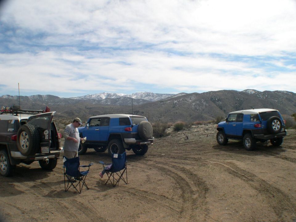 Three blue Toyota FJ Cruisers parked on a dirt track, with snow-capped mountains in the distance. Two camping chairs are set up