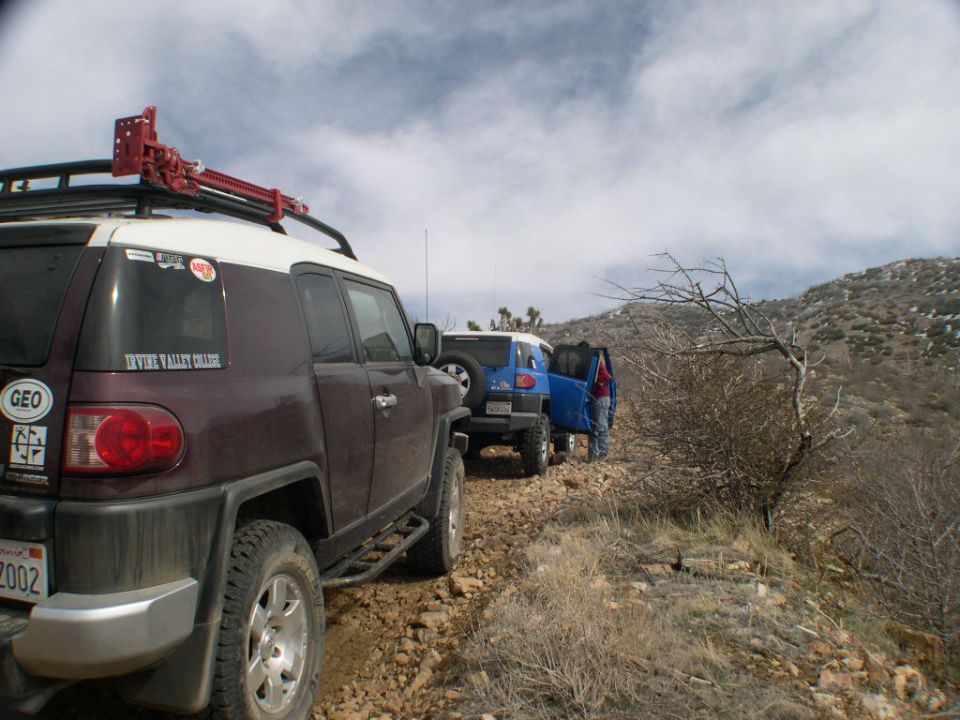 Dark red 2007 Toyota FJ Cruiser with roof rack and jack, parked on a rocky trail with another FJ Cruiser behind it