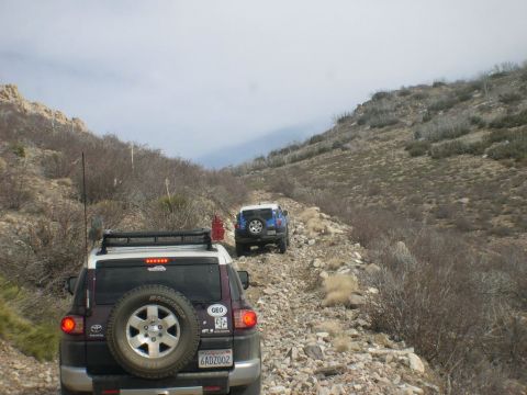 2007 Toyota FJ Cruiser - Two FJ Cruisers navigate a rough, mountainous off-road path under a cloudy sky.