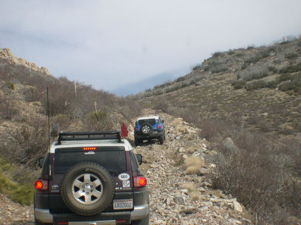 Two Toyota FJ Cruisers driving on a rocky, uphill trail with sparse vegetation