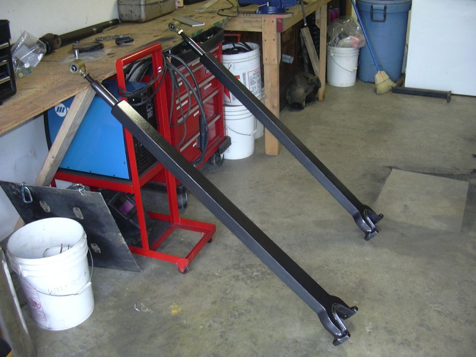 Pair of black metal radius arms with adjustable ends, lying on a concrete floor in a workshop