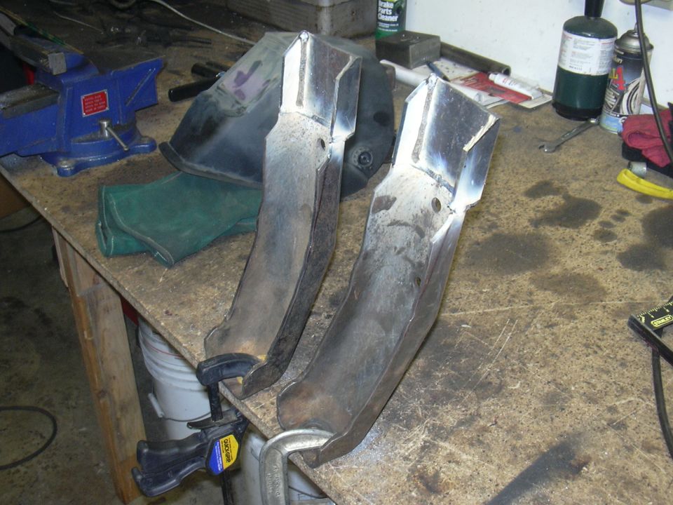 Two metal parts clamped to a workbench with a welding mask and vise in the background