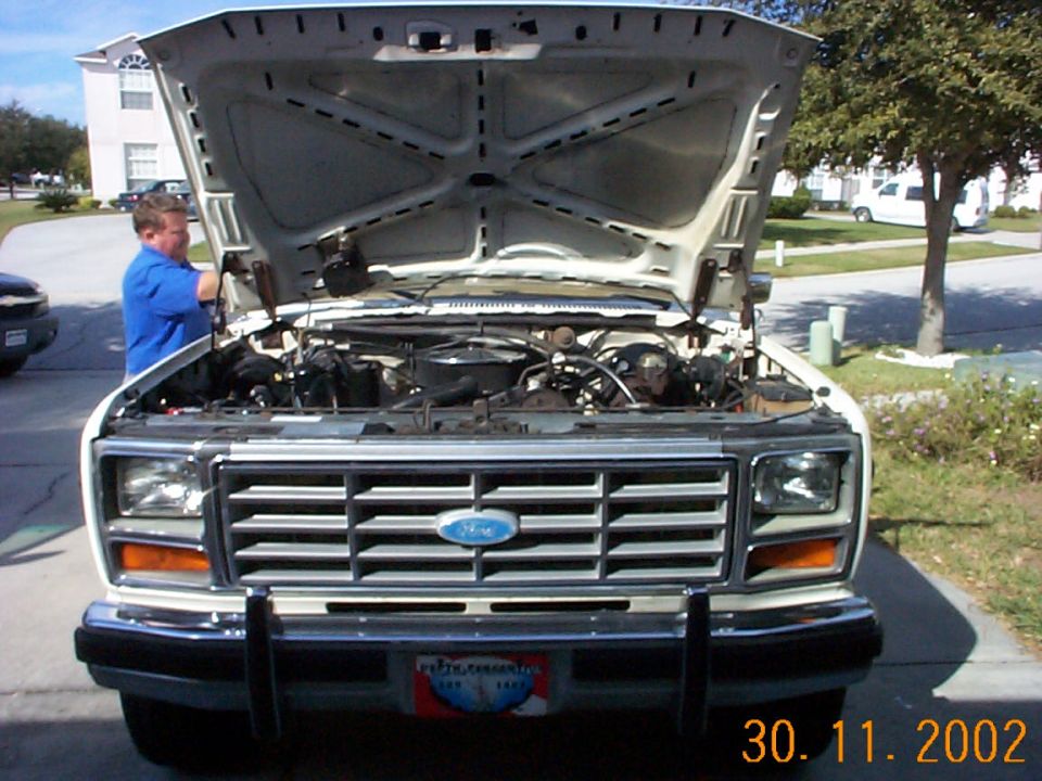 White 1980s Ford truck, hood up, man working on the engine in a driveway.