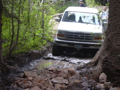1991 Ford Bronco - A white Ford van tackles a challenging muddy obstacle on a wooded trail.