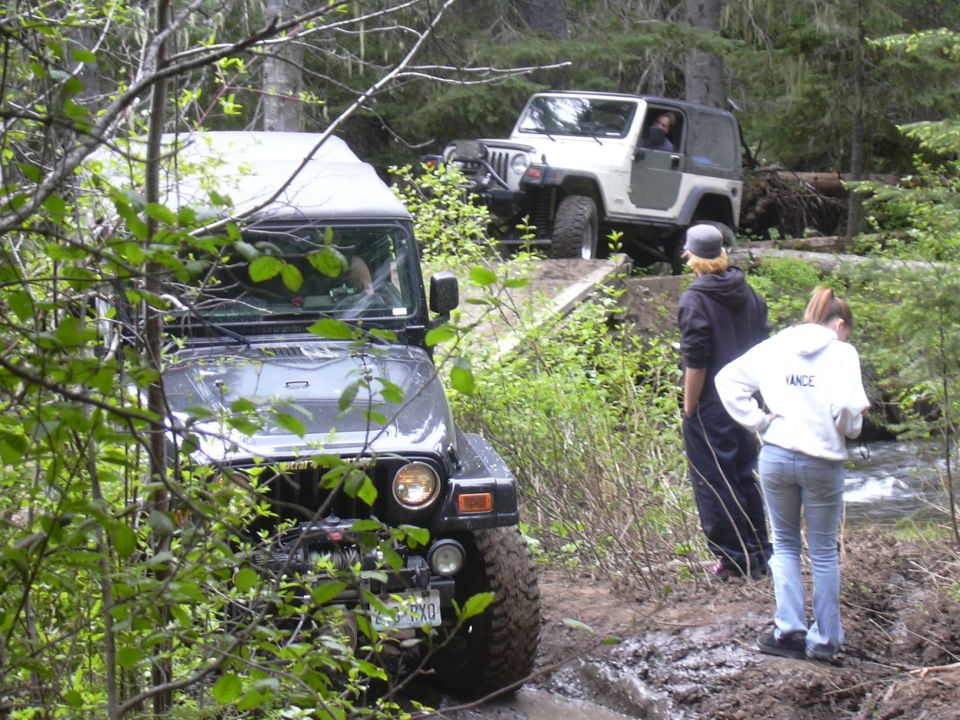 Two Jeep Wranglers on a muddy trail, one in front, one behind on a bridge. People watching