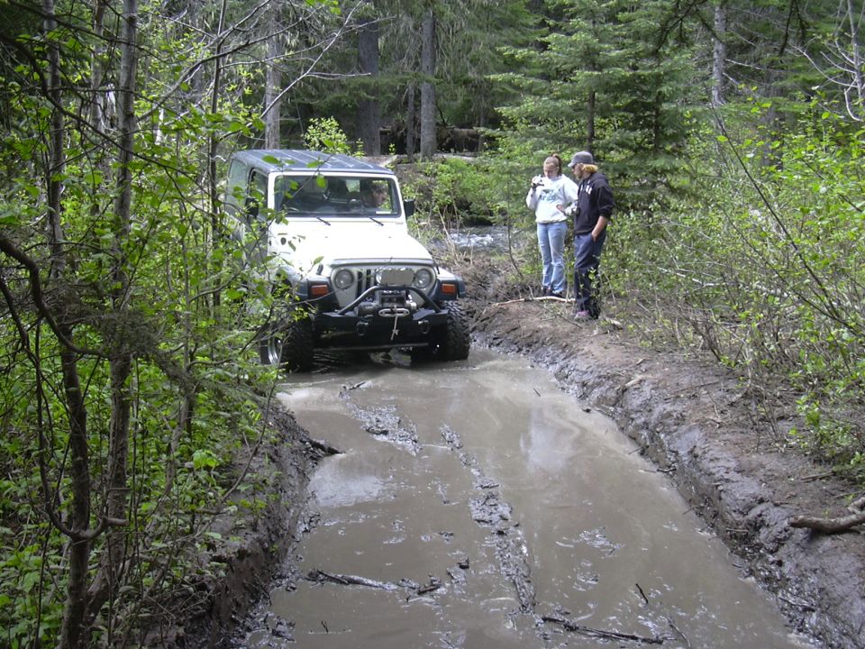 White Jeep Wrangler driving through a muddy forest trail with two people watching
