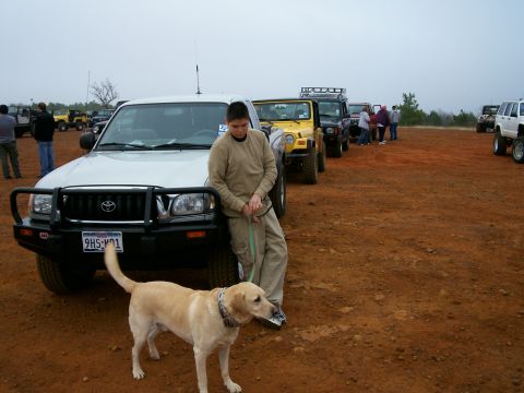 1993 Ford Bronco - A group of off-road vehicles are gathered on a dirt lot, with a boy and his dog standing by a Toyota Tacoma.