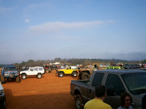 1993 Ford Bronco - A collection of modified off-road vehicles are gathered on a dirt field, with a clear blue sky overhead.