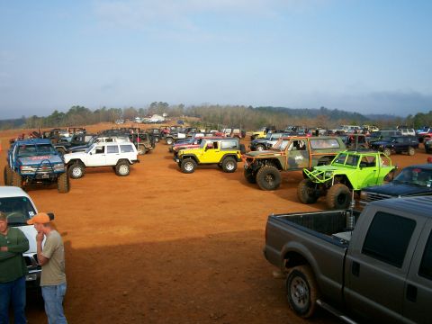 1993 Ford Bronco - A variety of off-road vehicles are parked on a dirt lot, with trees and hills in the background under a clear sky.