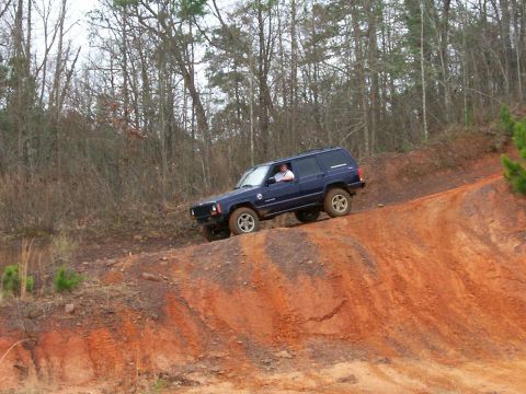 1993 Ford Bronco - A driver navigates a dark blue Jeep Cherokee XJ up a steep, red dirt incline in a wooded area.