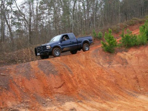 1993 Ford Bronco - A dark blue pickup truck navigates a steep, red dirt incline surrounded by trees.