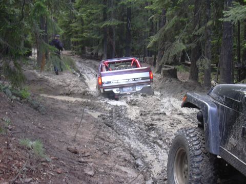 1991 Ford Bronco - A purple pickup truck with a roll cage tackles a challenging muddy trail, with another off-road vehicle visible in the foreground.