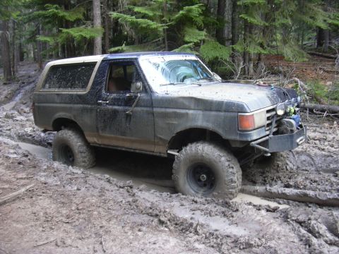 1991 Ford Bronco - A muddy 1991 Ford Bronco navigates a challenging off-road trail, its tires deep in the mire.