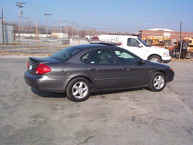 Dark gray Ford Taurus sedan, passenger-side rear 3/4 view in an industrial lot