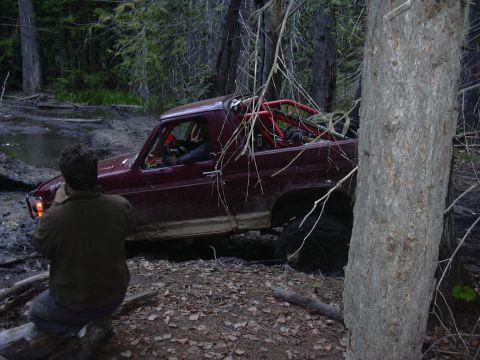 1991 Ford Bronco - A maroon off-road truck with a red roll cage is stuck in the mud, with a person observing from the side.