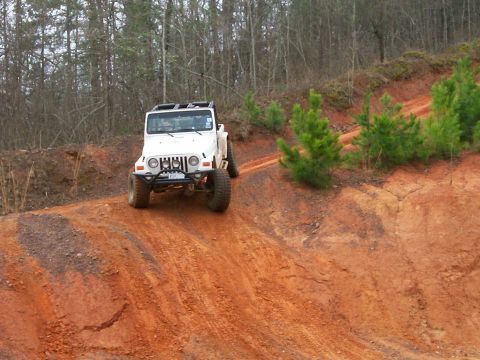 1993 Ford Bronco - A white Jeep Wrangler navigates a steep, red dirt trail surrounded by trees.