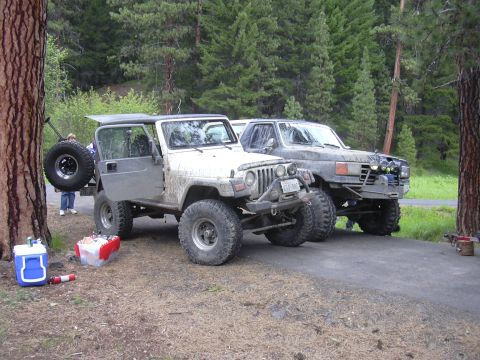 1991 Ford Bronco - Two off-road vehicles are parked on a paved surface next to a wooded area.