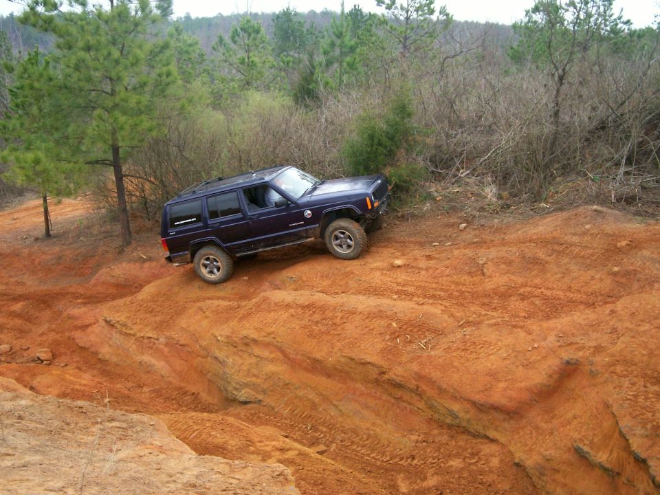 Dark blue Jeep Cherokee XJ driving on a red dirt trail with trees in the background