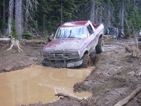 1991 Ford Bronco - This lifted Ford truck is deep in a mud hole on an off-road trail, with other vehicles and people nearby.