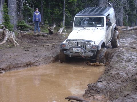 1991 Ford Bronco - A white Jeep Wrangler tackles a muddy obstacle on an off-road trail, with a person watching from the side.
