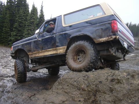 1991 Ford Bronco - A driver maneuvers a dark blue Ford Bronco through a challenging off-road course with muddy terrain and trees in the background.