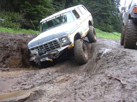 1991 Ford Bronco - White Ford Bronco navigates a muddy off-road trail, with another vehicle visible behind it.