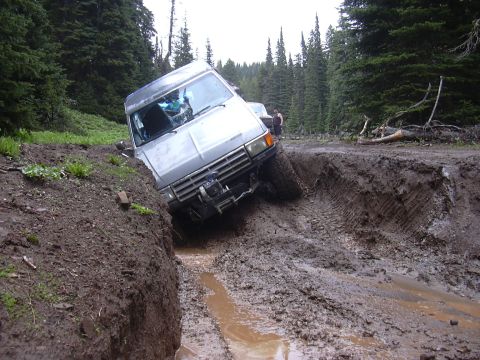 1991 Ford Bronco - A silver 1991 Ford Bronco is stuck in a deep muddy ditch on a forest road, with trees in the background.
