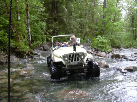 1991 Ford Bronco - A white Jeep CJ-5 navigates a shallow, rocky riverbed surrounded by trees.