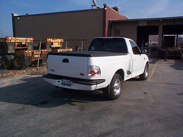 White regular-cab Ford F-150 with tonneau cover, 3/4 rear view in a shop parking lot