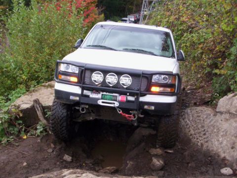 1991 Ford Bronco - A white SUV with a winch and off-road tires is shown navigating a challenging rocky and muddy path.