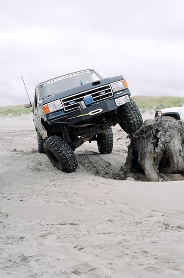 1991 Ford Bronco - 1991 Ford Bronco tackles a sand dune obstacle with driftwood, another SUV in background.