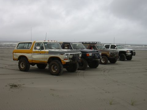 1991 Ford Bronco - Four modified trucks, including a 1978 Ford Bronco, are parked on a beach under an overcast sky.