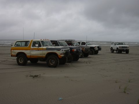 1991 Ford Bronco - A group of SUVs and trucks are parked on the sand near the ocean under a cloudy sky.