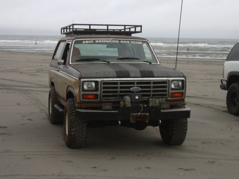 1991 Ford Bronco - Ford Bronco with roof rack and club sticker parked on a sandy beach near the ocean.