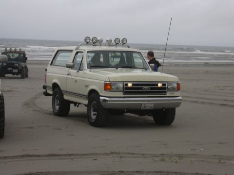 1991 Ford Bronco - A 1991 Ford Bronco with auxiliary lights on the roof drives along the beach, with another vehicle visible in the background.