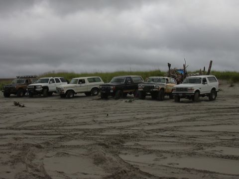 1991 Ford Bronco - Several SUVs, including a white Ford Bronco, are lined up on a sandy beach under a cloudy sky.