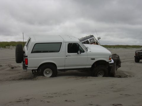 1991 Ford Bronco - A white Ford Bronco is stuck in the sand on a beach, with another vehicle partially visible behind it.