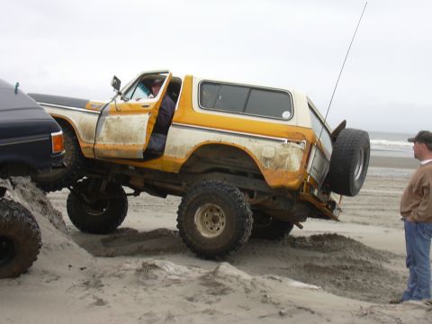 1991 Ford Bronco - Two off-road vehicles navigate a sandy beach with the ocean in the background.