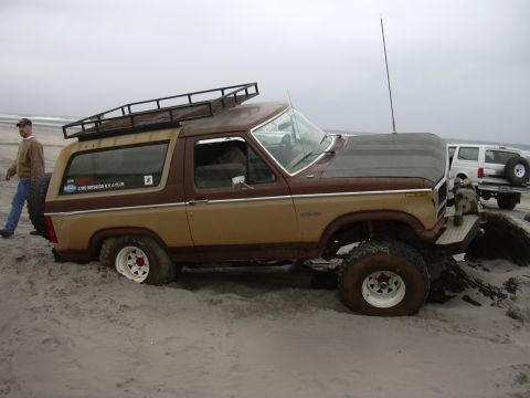 1991 Ford Bronco - A two-tone Ford Bronco is stuck in the sand on a beach, with another SUV visible in the background.
