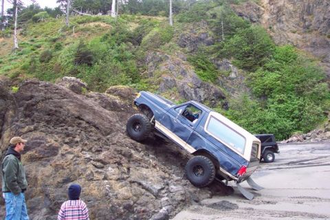 1991 Ford Bronco - A blue Ford Bronco navigates a rocky incline on a sandy beach, with another vehicle visible in the distance.