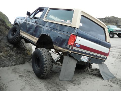 1991 Ford Bronco - A modified Ford Bronco navigates a rocky beach, with another lifted truck visible in the background.