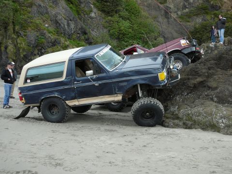 1991 Ford Bronco - Two off-road vehicles navigate a rocky beach with steep cliffs in the background.