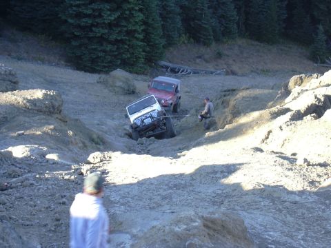 1991 Ford Bronco - Two off-road vehicles are stuck in a muddy, rocky ditch with people observing the situation.