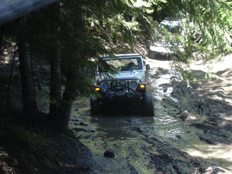 1991 Ford Bronco - A white Jeep Wrangler tackles a challenging muddy off-road path, with another vehicle visible in the background.