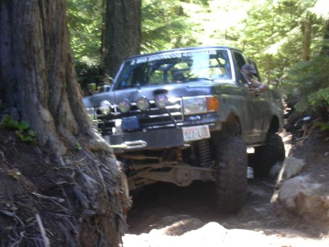 1991 Ford Bronco - A 1991 Ford Bronco navigates a narrow, wooded trail with large tires and auxiliary lights.