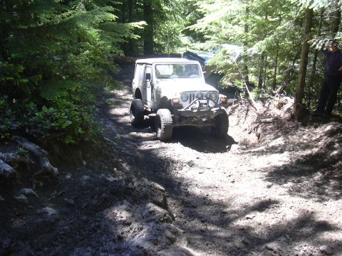 1991 Ford Bronco - A white Jeep Wrangler navigates a challenging muddy trail through a dense forest.