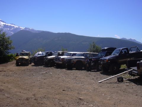 1991 Ford Bronco - A line of off-road vehicles are parked on a dirt lot with a mountain range visible in the distance under a clear blue sky.