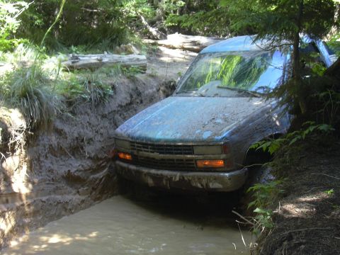 1991 Ford Bronco - A blue pickup truck navigates a challenging muddy trail, getting stuck in a deep rut.