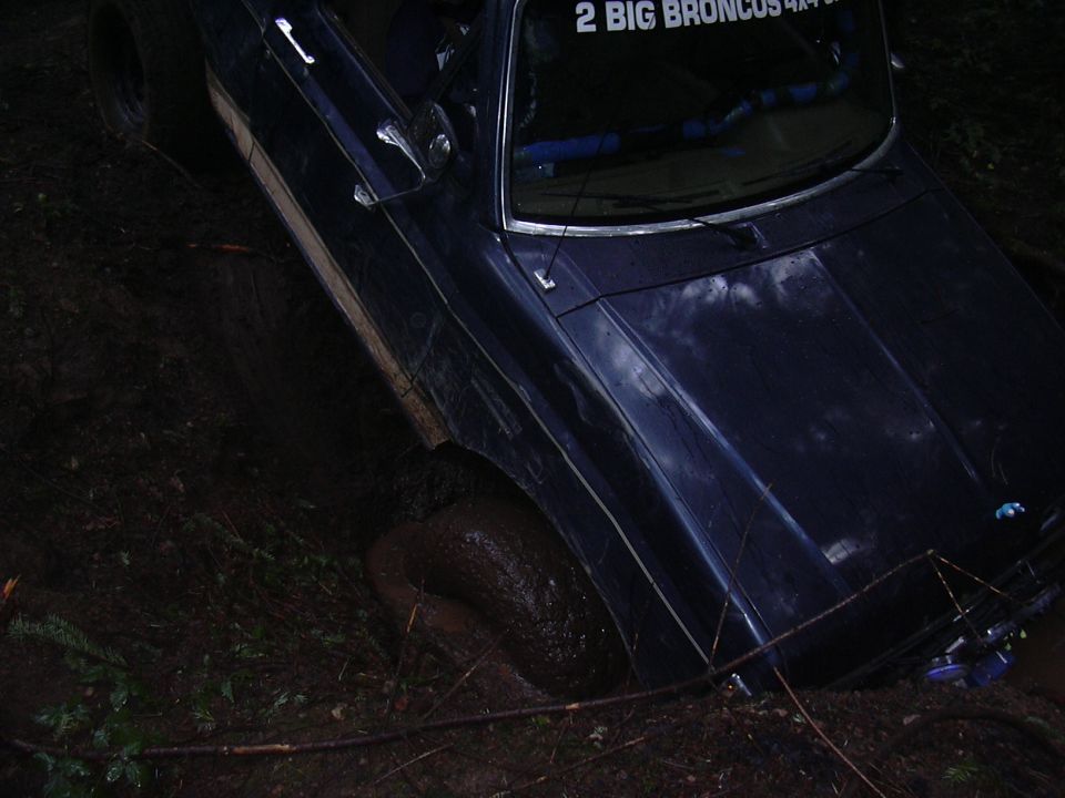 Dark blue SUV stuck in mud, front right wheel submerged. Sticker on rear window reads '2 BIG BRONCOS 4X4'
