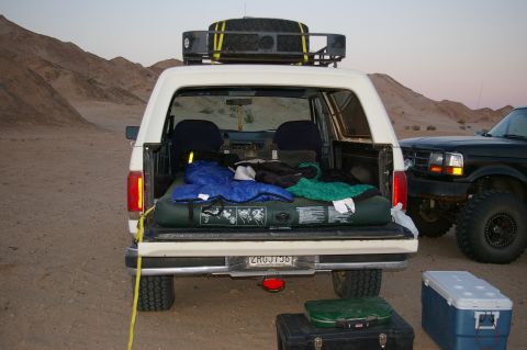 1986 Ford Bronco - A white Ford Bronco is parked in a desert landscape with camping gear loaded in its open rear, next to a black Ford truck.